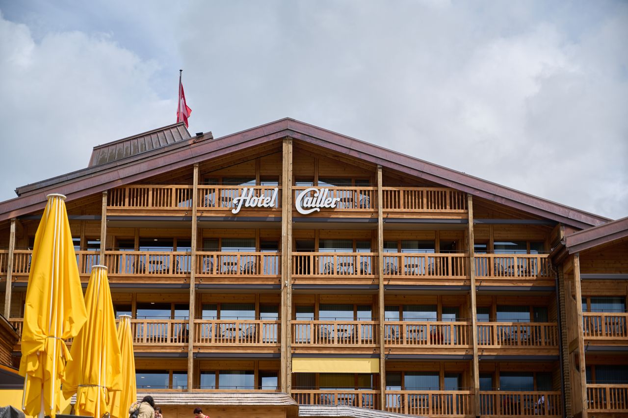 about-img Rustic wooden architecture of Hotel Cailler with balconies and yellow umbrellas in Charmey, Switzerland.
